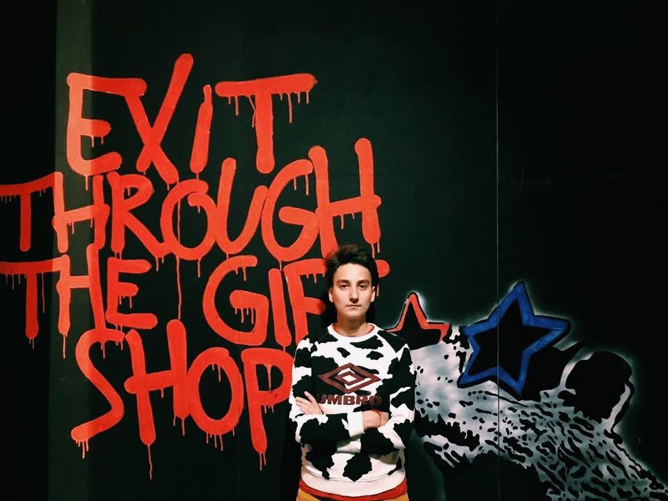 Maria Zhukova standing arms crossed in front of red graffiti that reads Exit through the gift shop